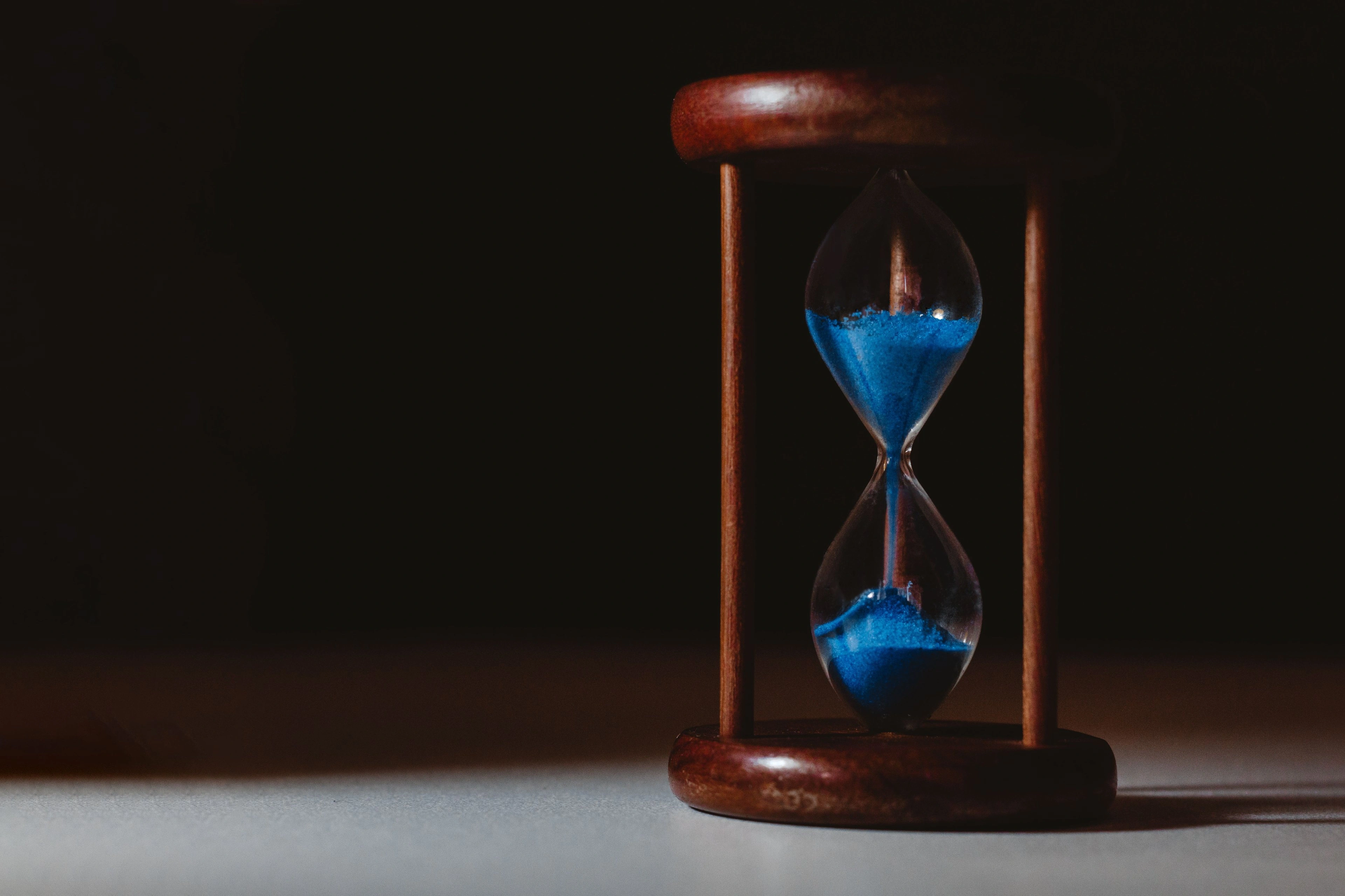 An hourglass with blue sand on a dark surface, lit low against deep shadow.