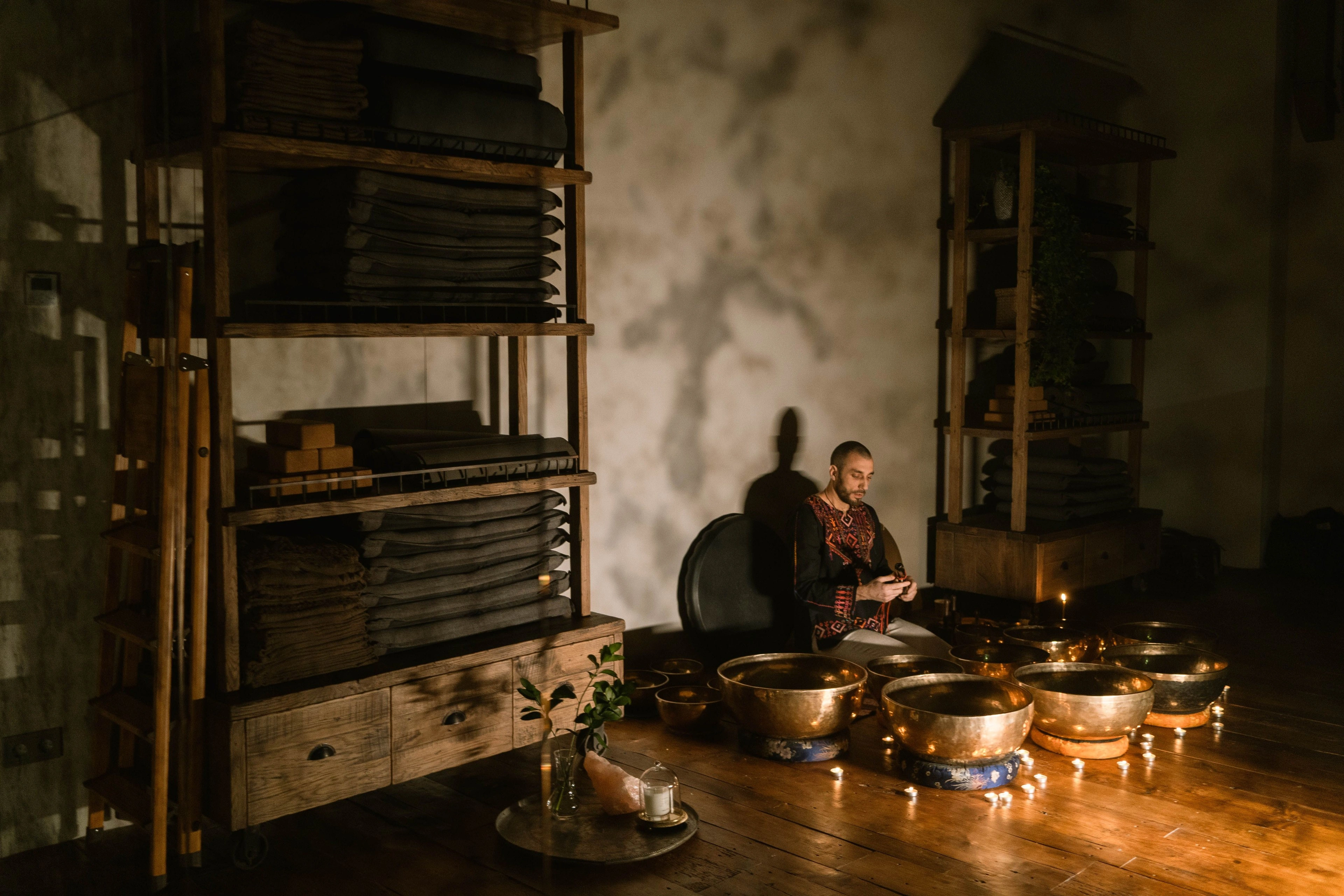 A person seated among brass singing bowls in a dim, warmly lit room.