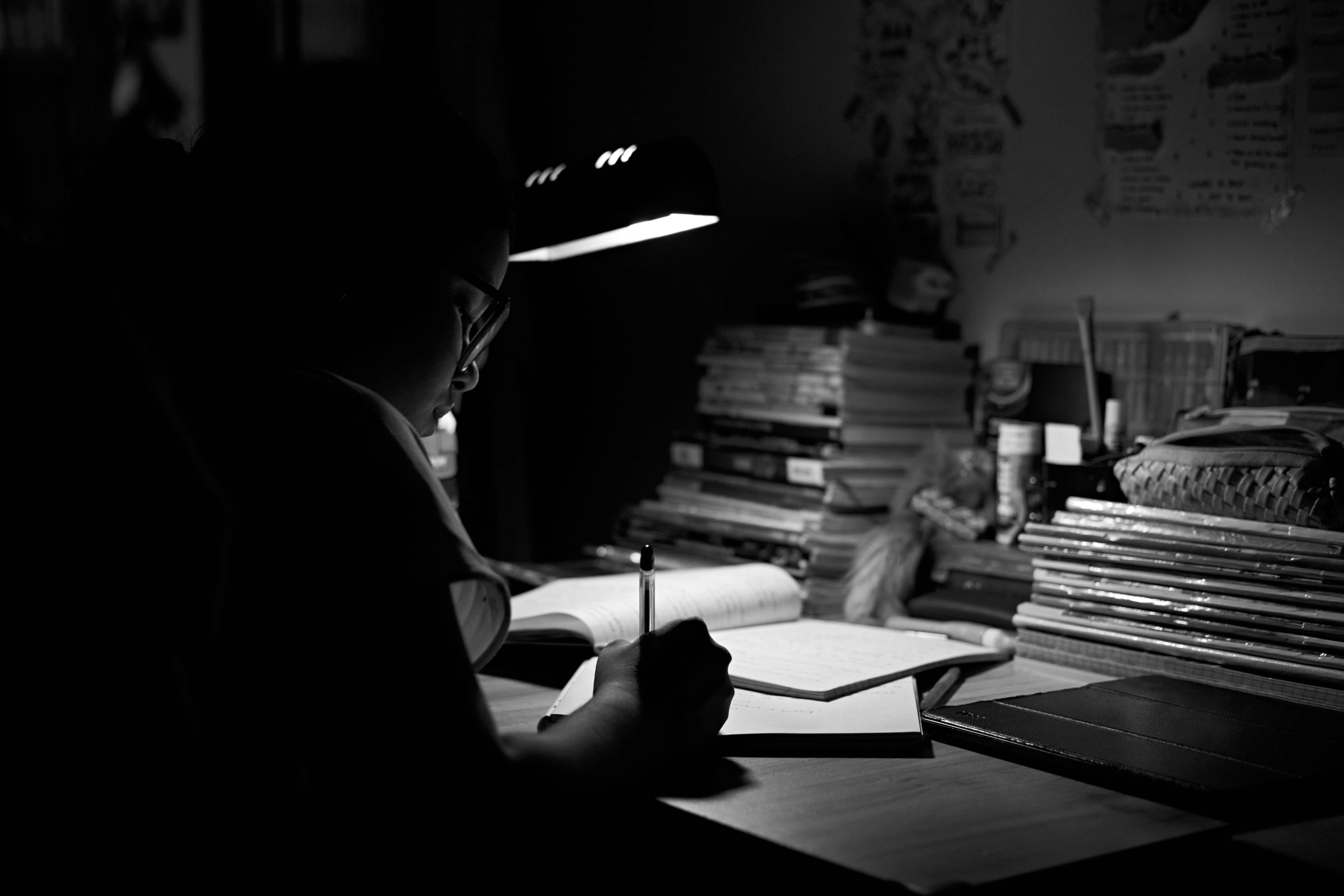 A dim desk lit by a single lamp, books and papers falling into shadow.