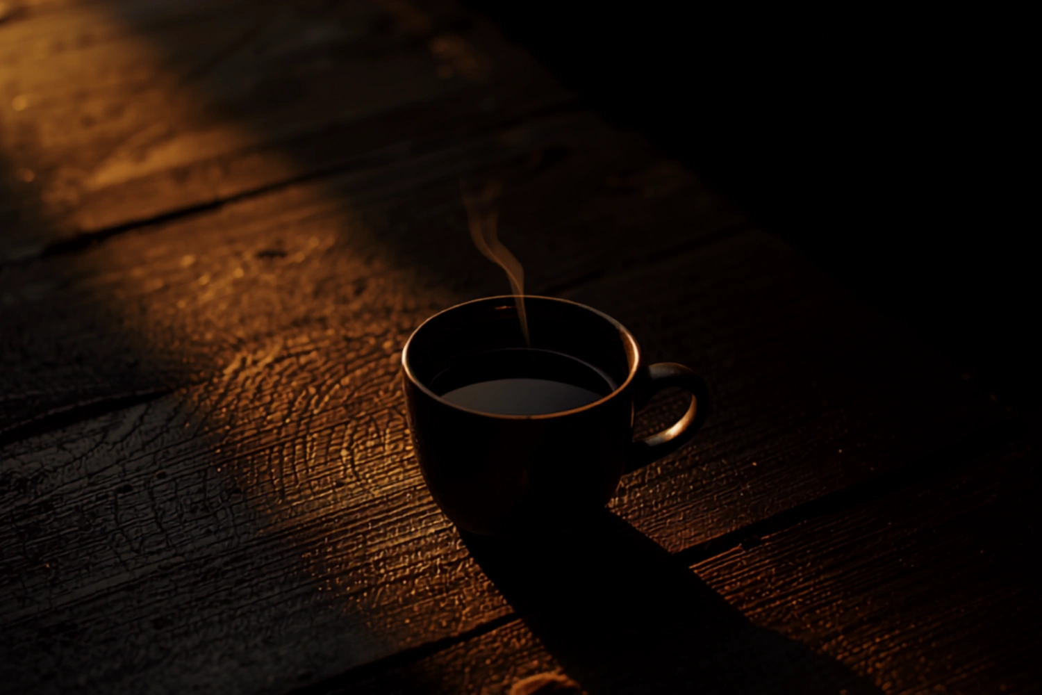 A dark ceramic cup of black coffee on a weathered wooden surface, steam catching a single beam of warm gold light against deep shadow.