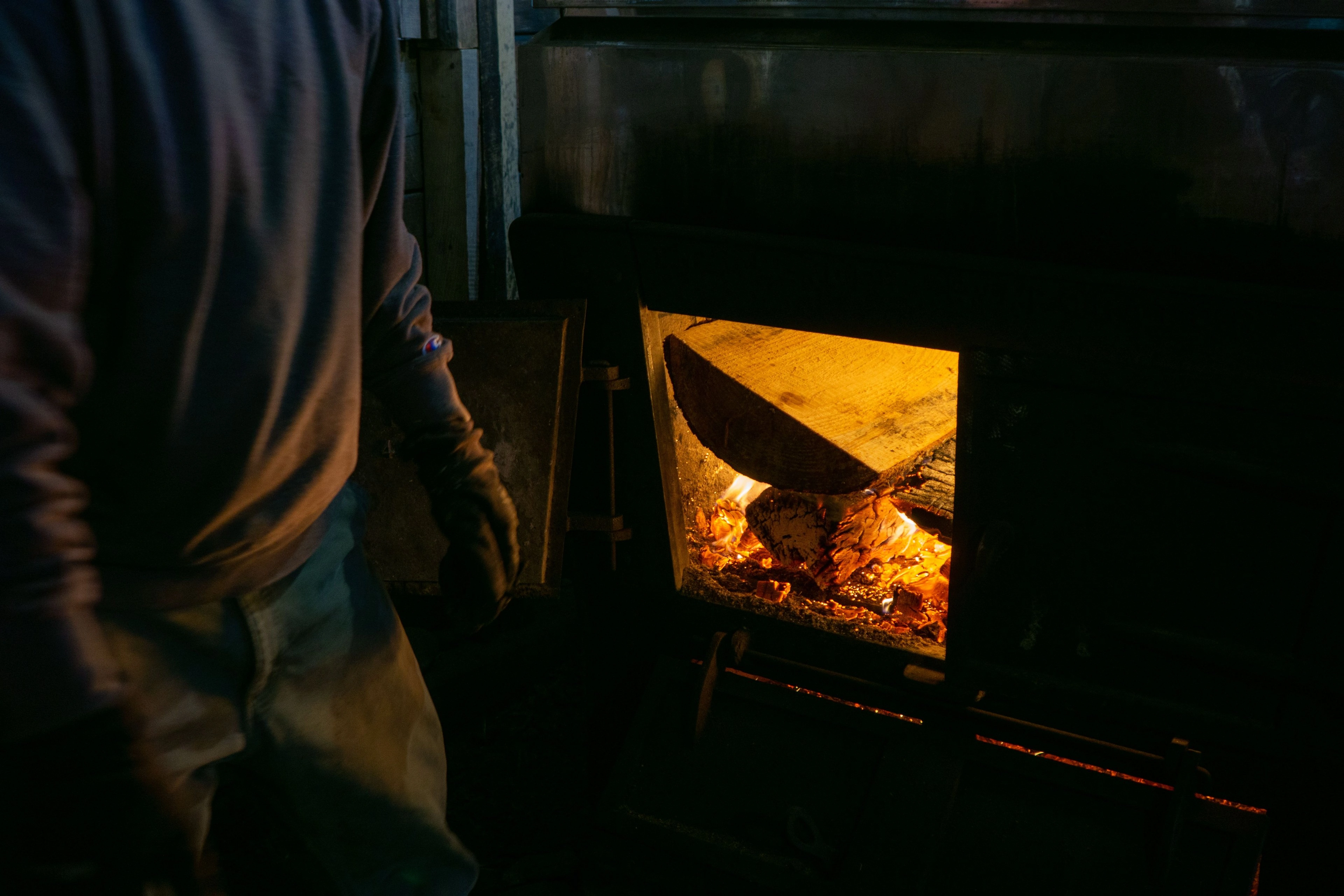 A figure tending a glowing fire in a dark hearth.