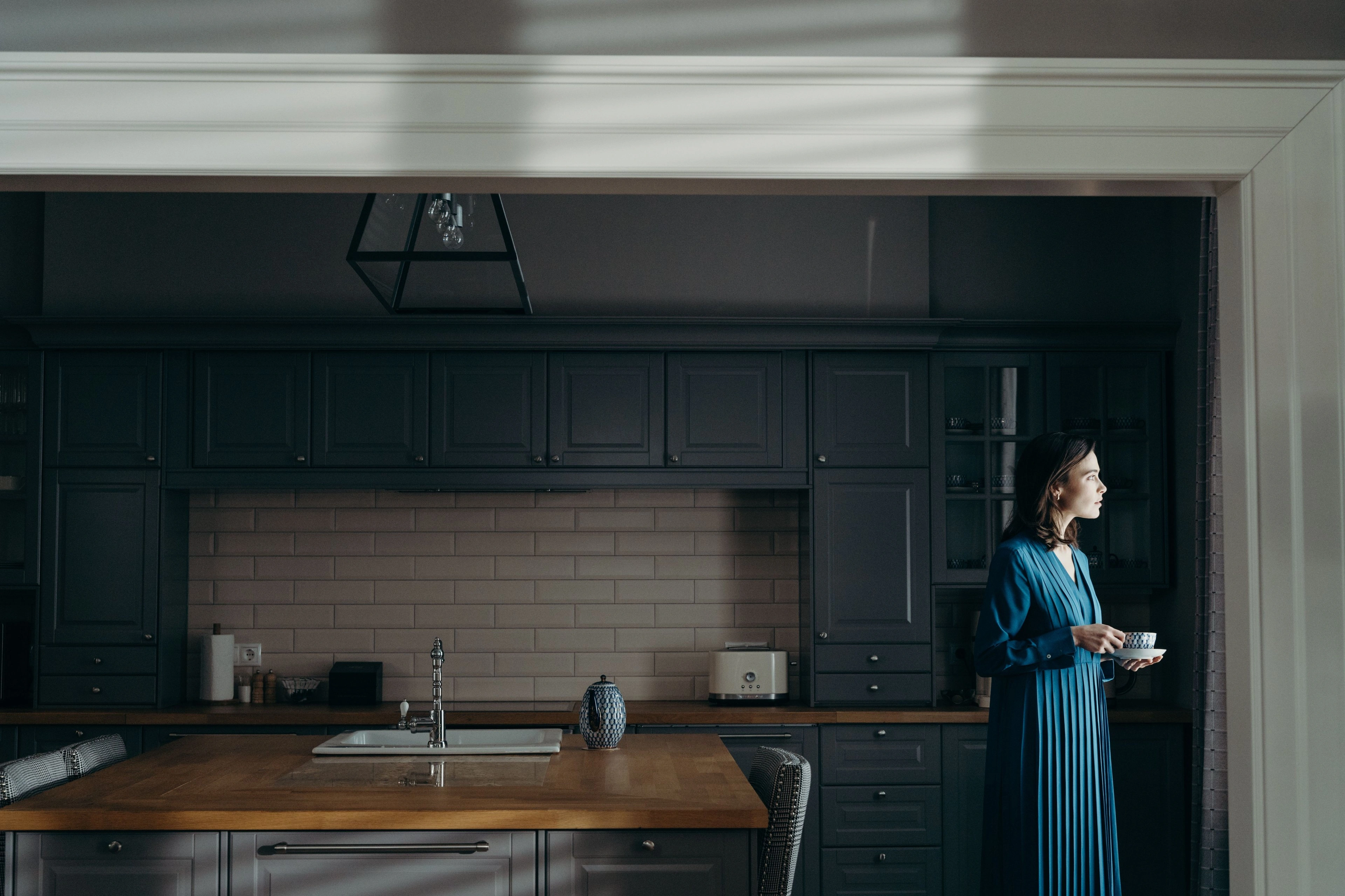 A figure standing with a cup in a dim, quiet kitchen.