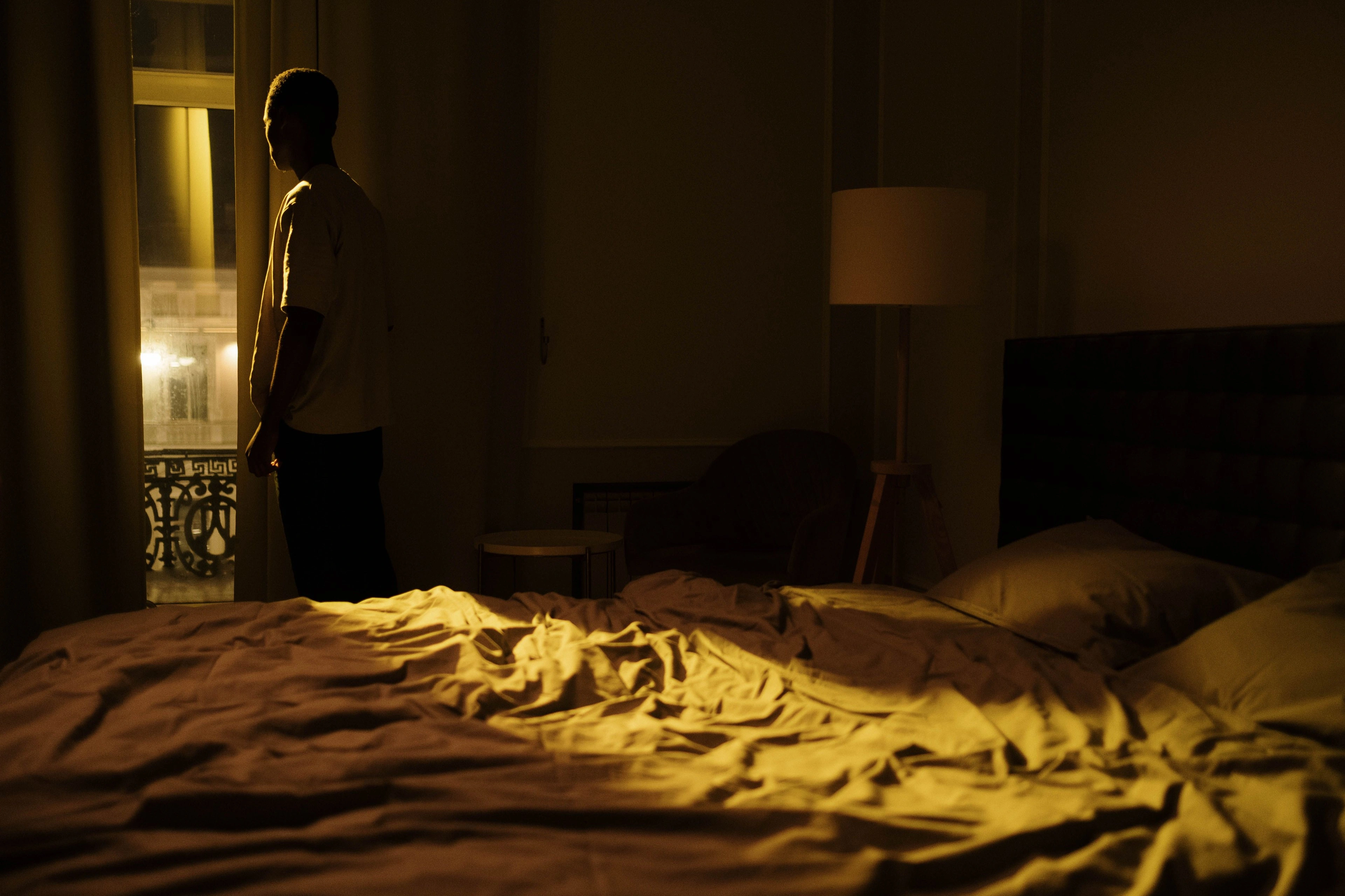 A dark bedroom with rumpled bedding and a figure by a lit doorway.
