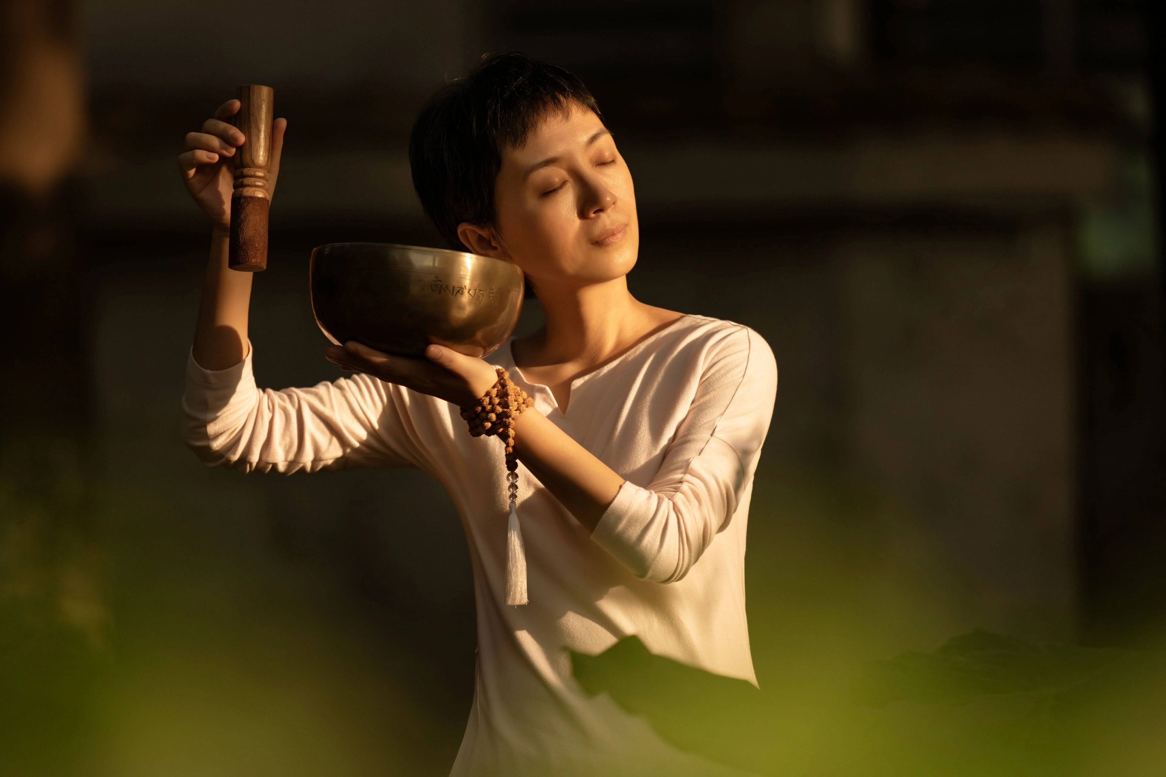 A woman holding a brass singing bowl to her ear in warm golden light.