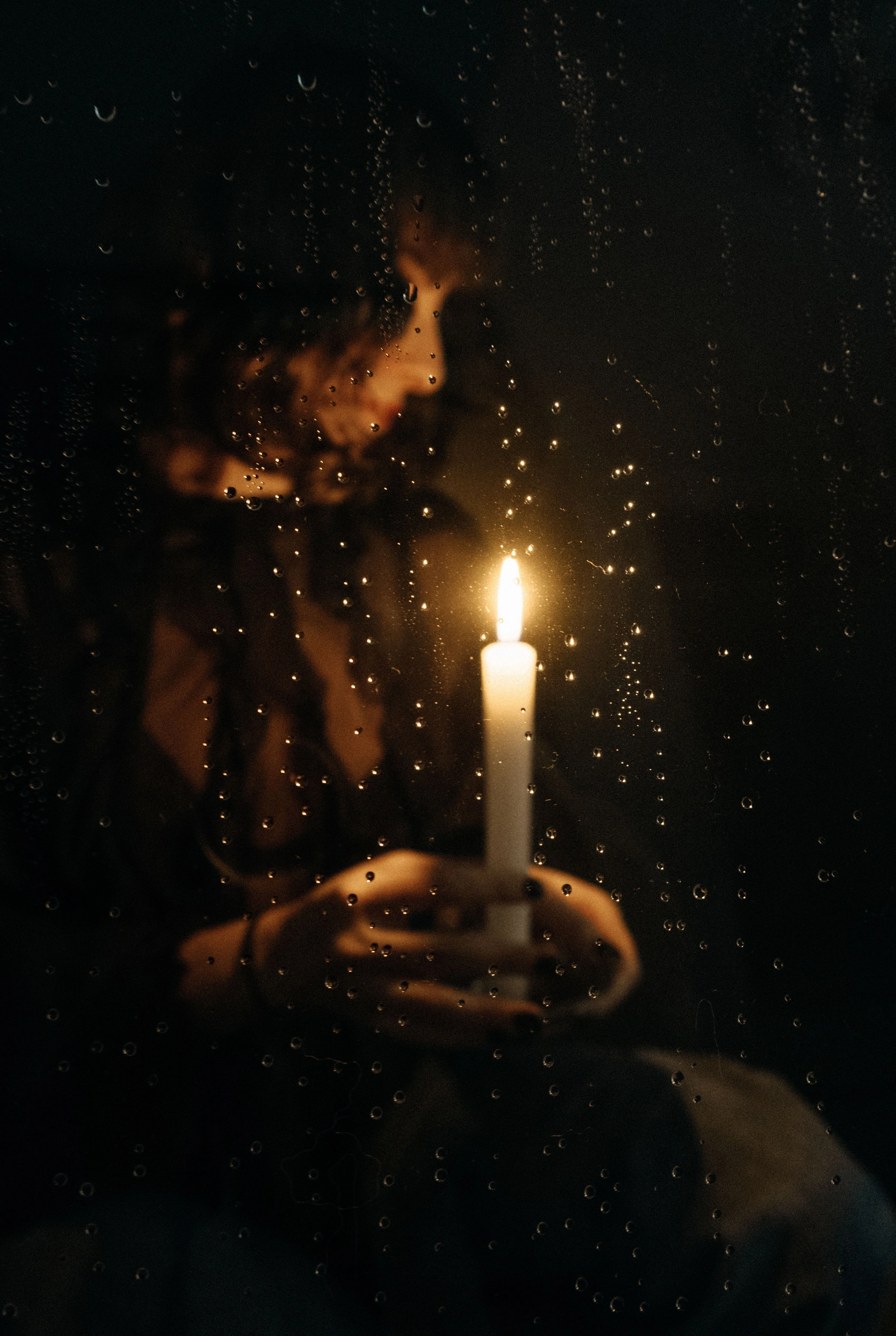 A candle held in cupped hands before a rain-streaked window.