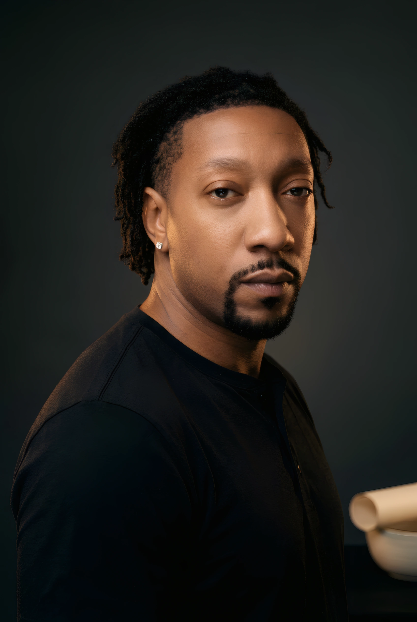Studio portrait of The Navigator, turned toward the camera in a black henley against a dark grey backdrop, a ceramic bowl resting on the table beside him.