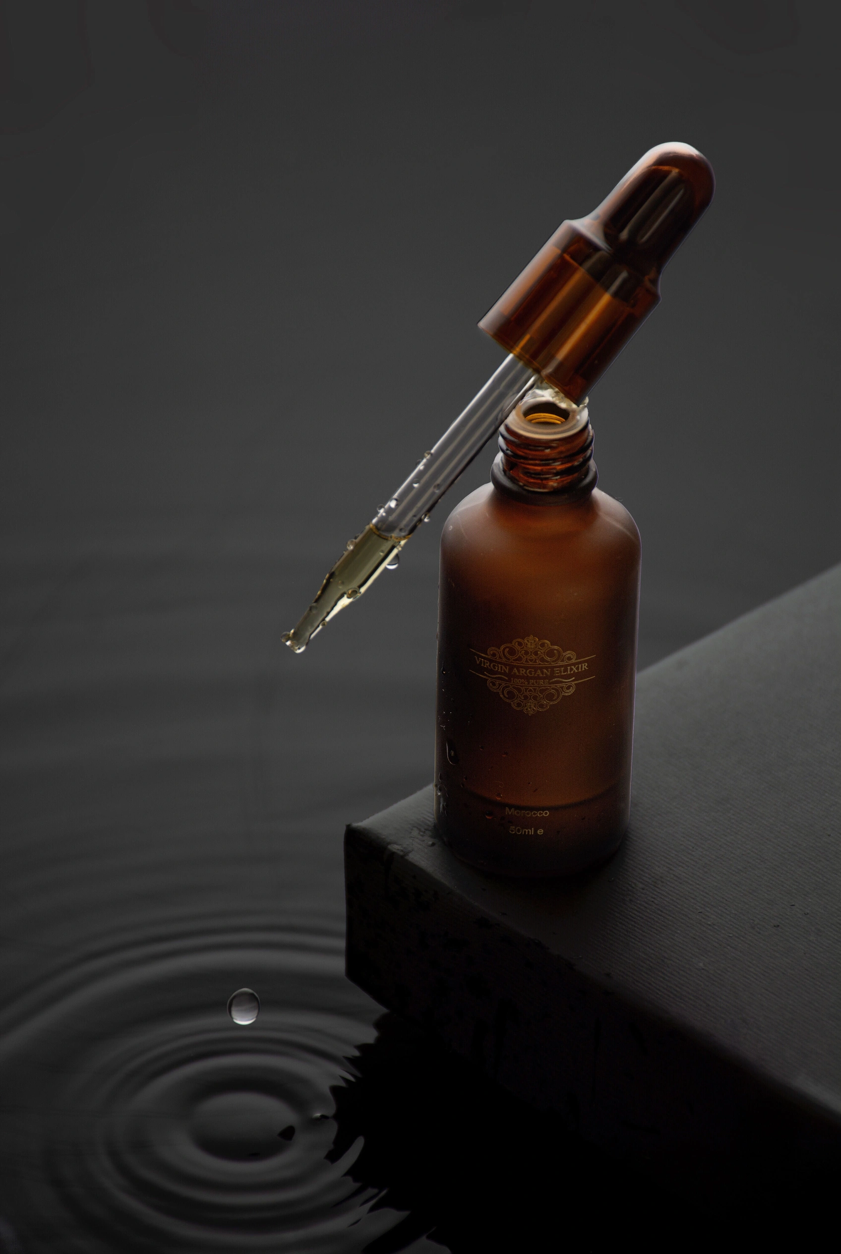 An amber dropper bottle on dark stone above a single ripple.