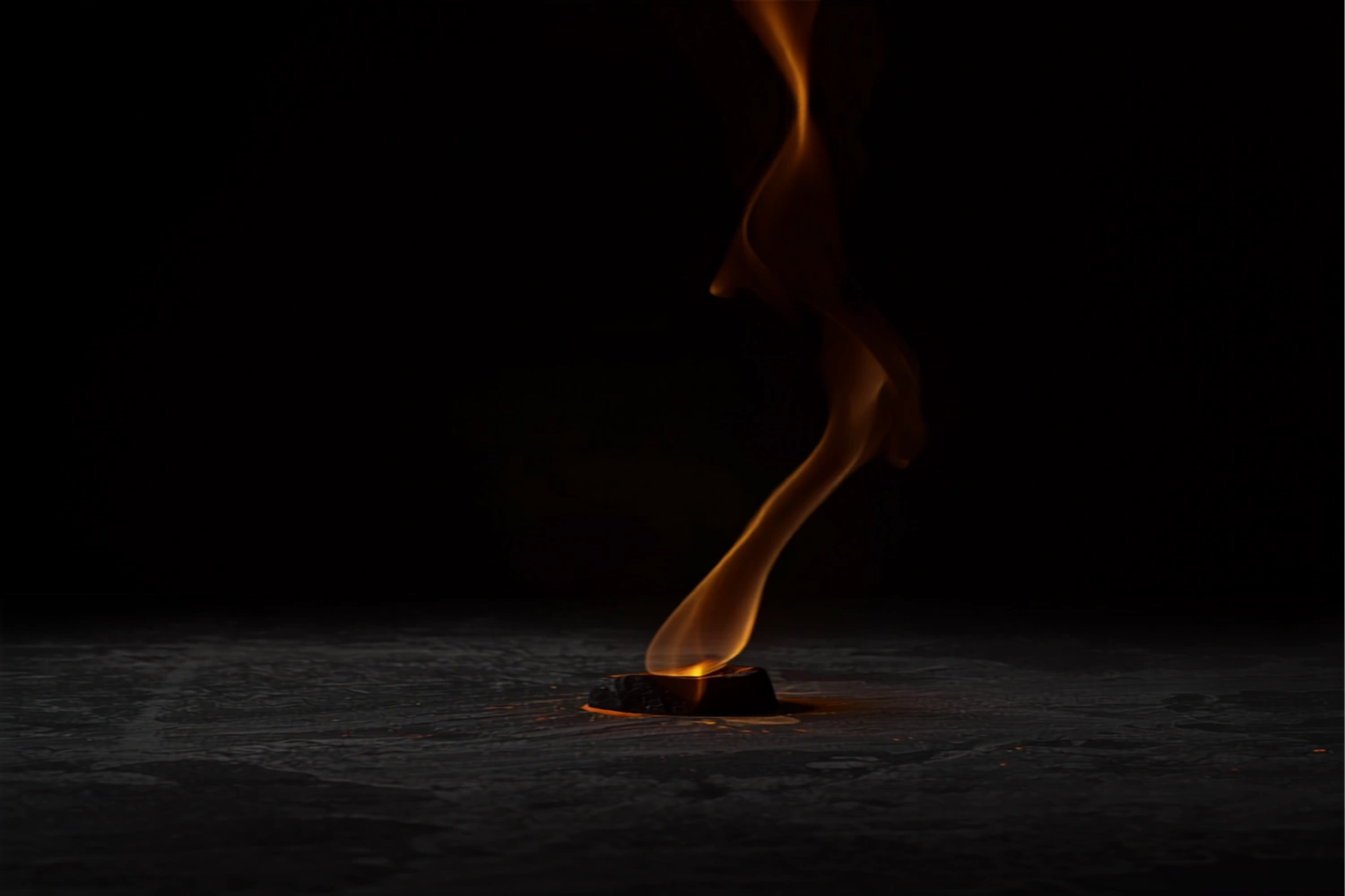 A single glowing ember rests on dark stone as one warm gold plume of smoke rises through a quiet, unlit background.