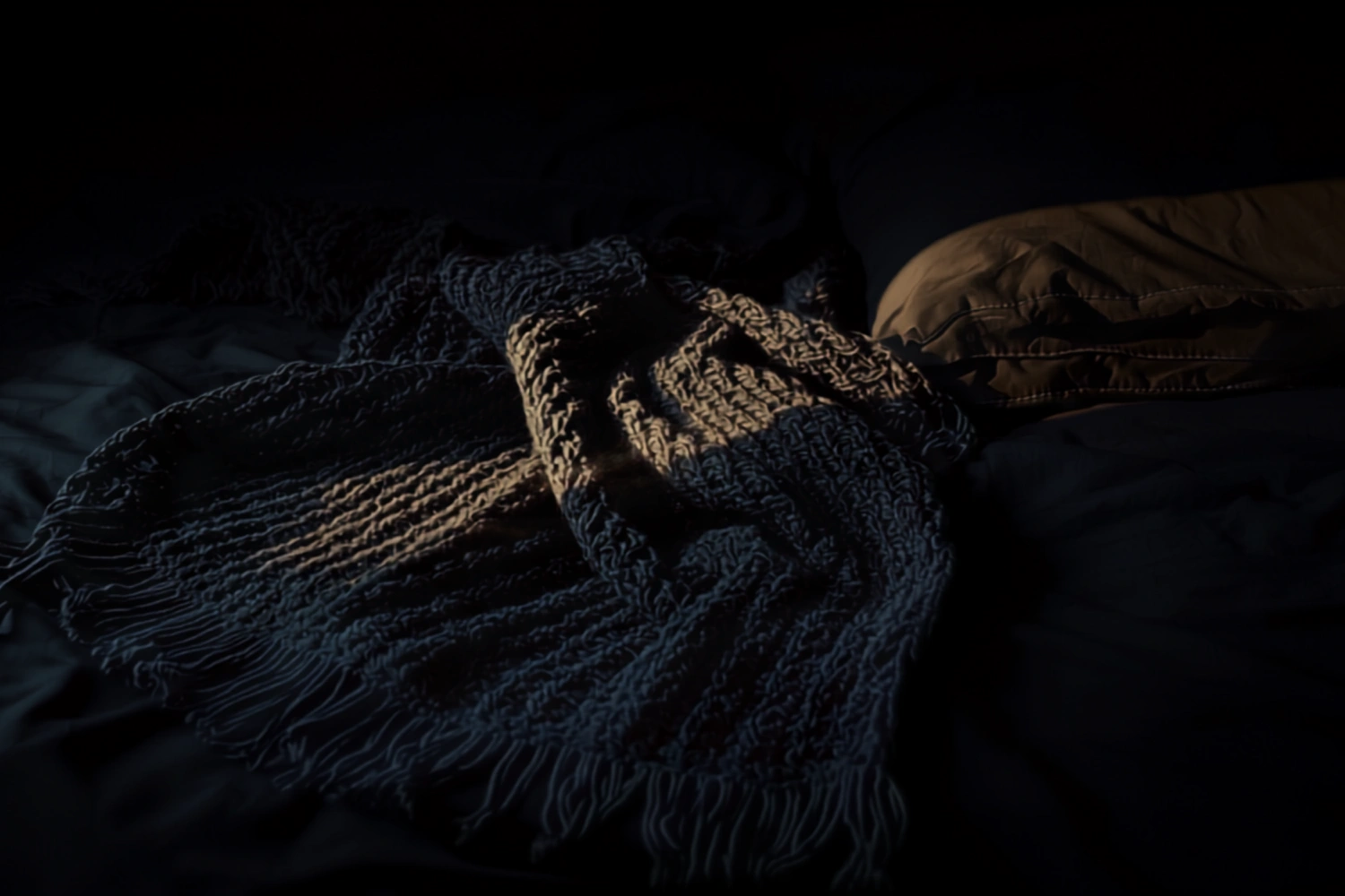 A heavy charcoal knit blanket folded on a dark bed, low warm gold light raking across the texture against deep shadow.